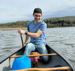 A young man with a baseball cap blue t shirt and jeans paddling a canoe. A small brown dog is with him in the bottom of the canoe