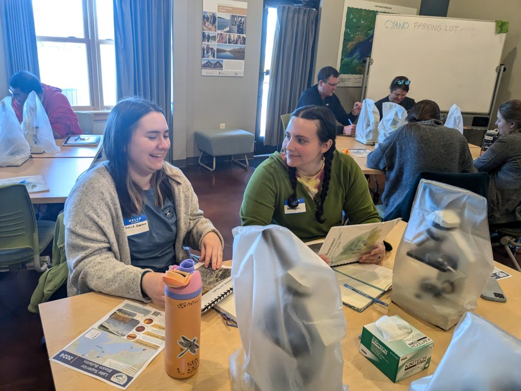 Two workshop attendees seated at a classroom table with workbooks and microscopes in front of them. They are laughing. More attendees are visible in the background.