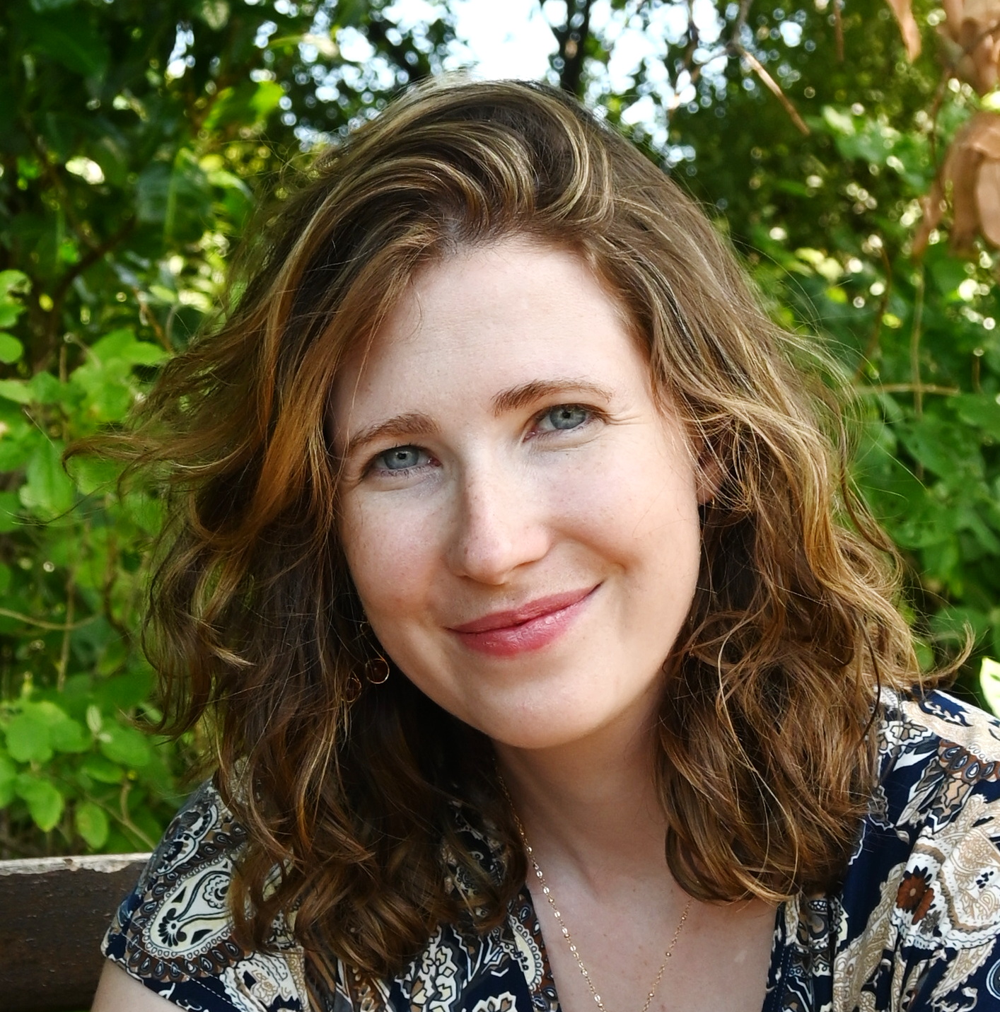 A close shot of a woman with brown curly hair smiling in front of a background of foliage